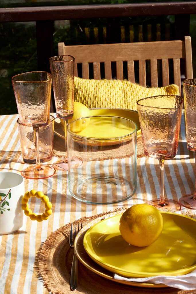 A cozy summer table on a veranda with lemons, yellow tableware, and textured glassware. Warm sunlight creates a cheerful, rustic, and welcoming atmosphere for an outdoor meal